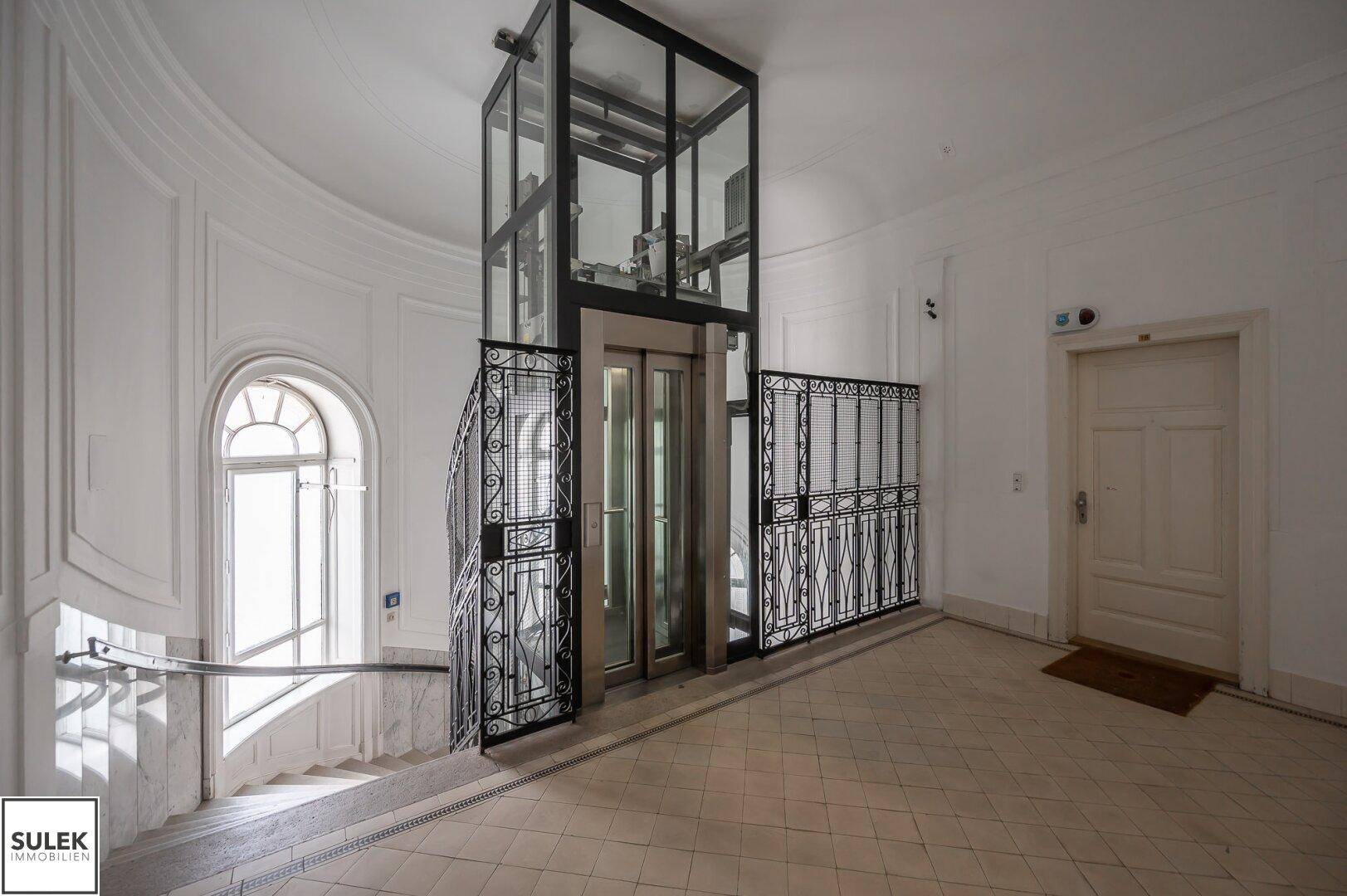 Heller Treppenaufgang mit modernem Glasaufzug und historischen Geländern.