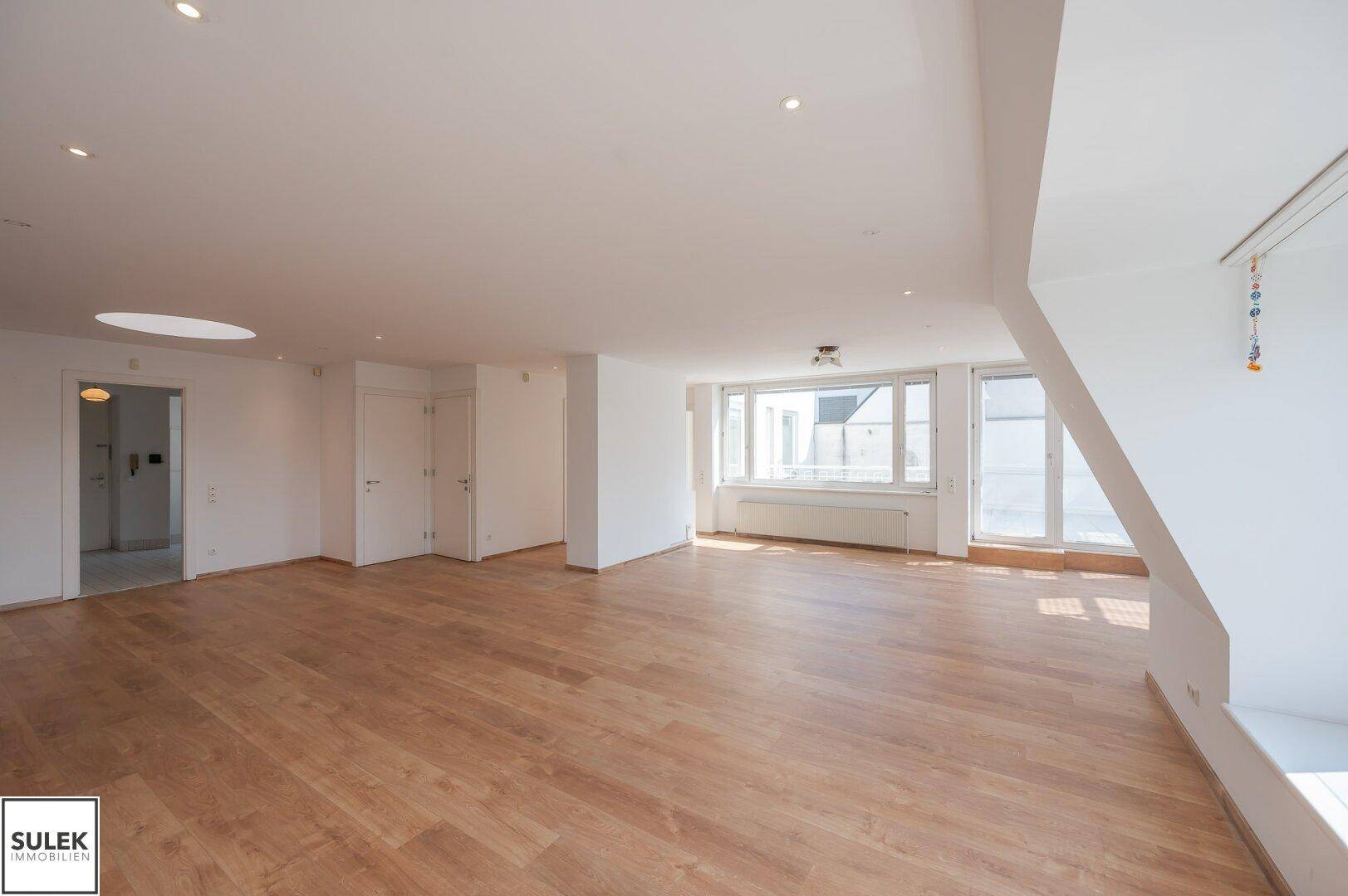 Offener Wohnbereich mit Parkettboden, Dachschrägen und viel Tageslicht durch große Fenster.