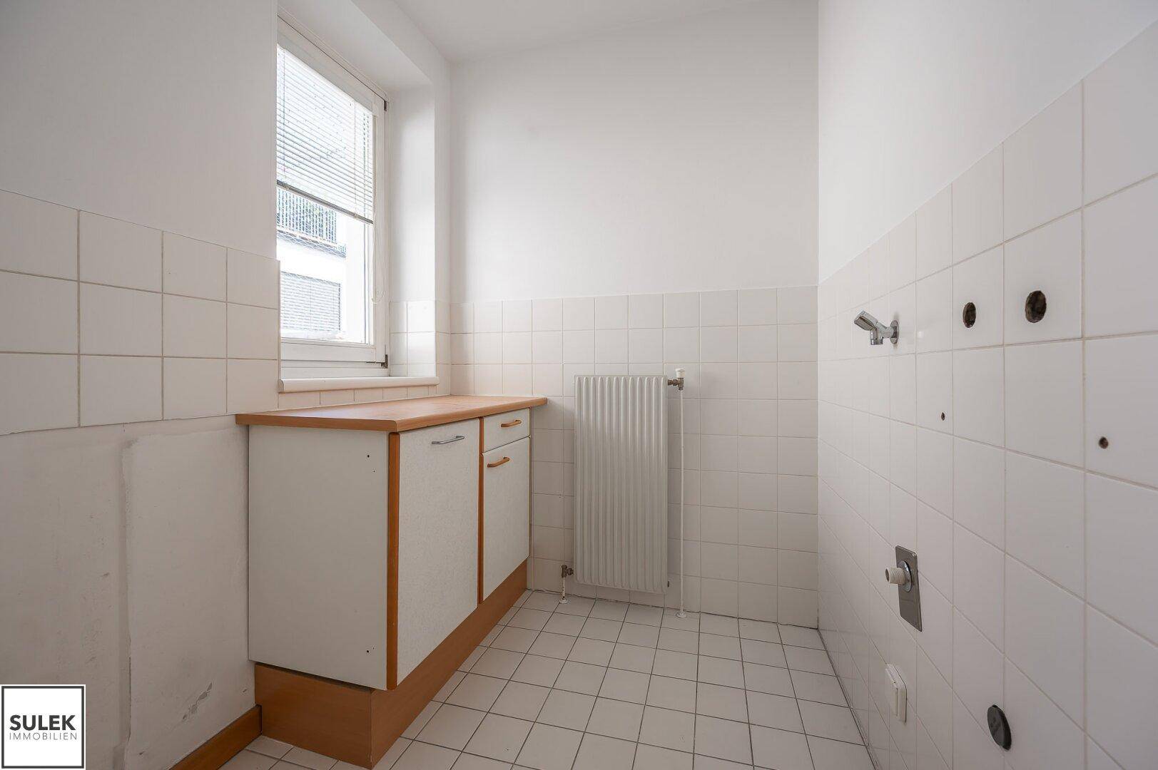 Kleiner Raum mit Fliesenboden, Fenster und Anschlüssen für Waschmaschine oder Spüle.