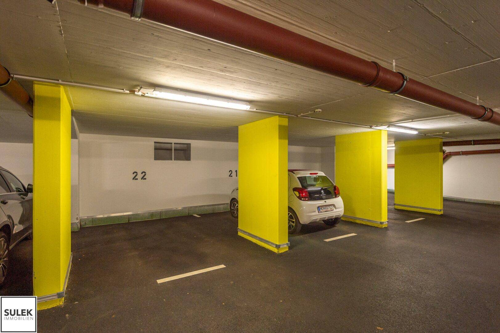 Helle Tiefgarage mit nummerierten Parkplätzen und sichtbaren Versorgungsleitungen an der Decke.