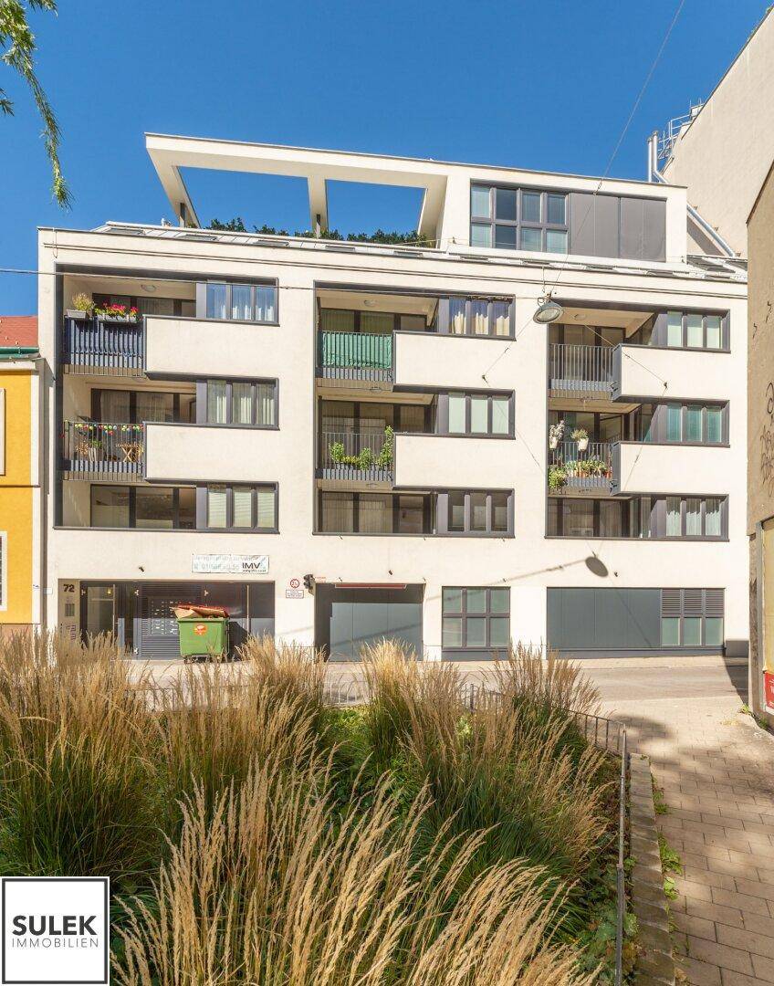 Moderne Wohnhausfassade mit Balkonen und Dachterrasse, umgeben von städtischer Vegetation.