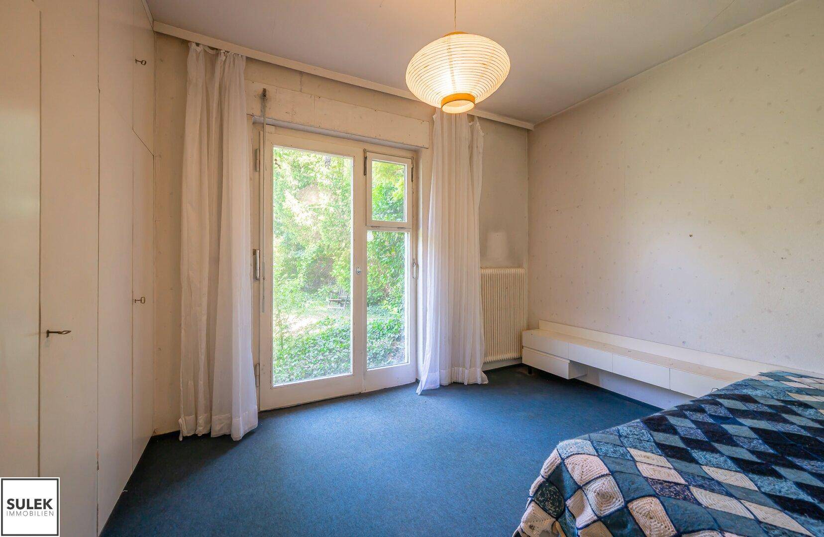 Ein Schlafzimmer mit blauem Teppichboden und einer großen Fenstertür mit Blick ins Grüne.
