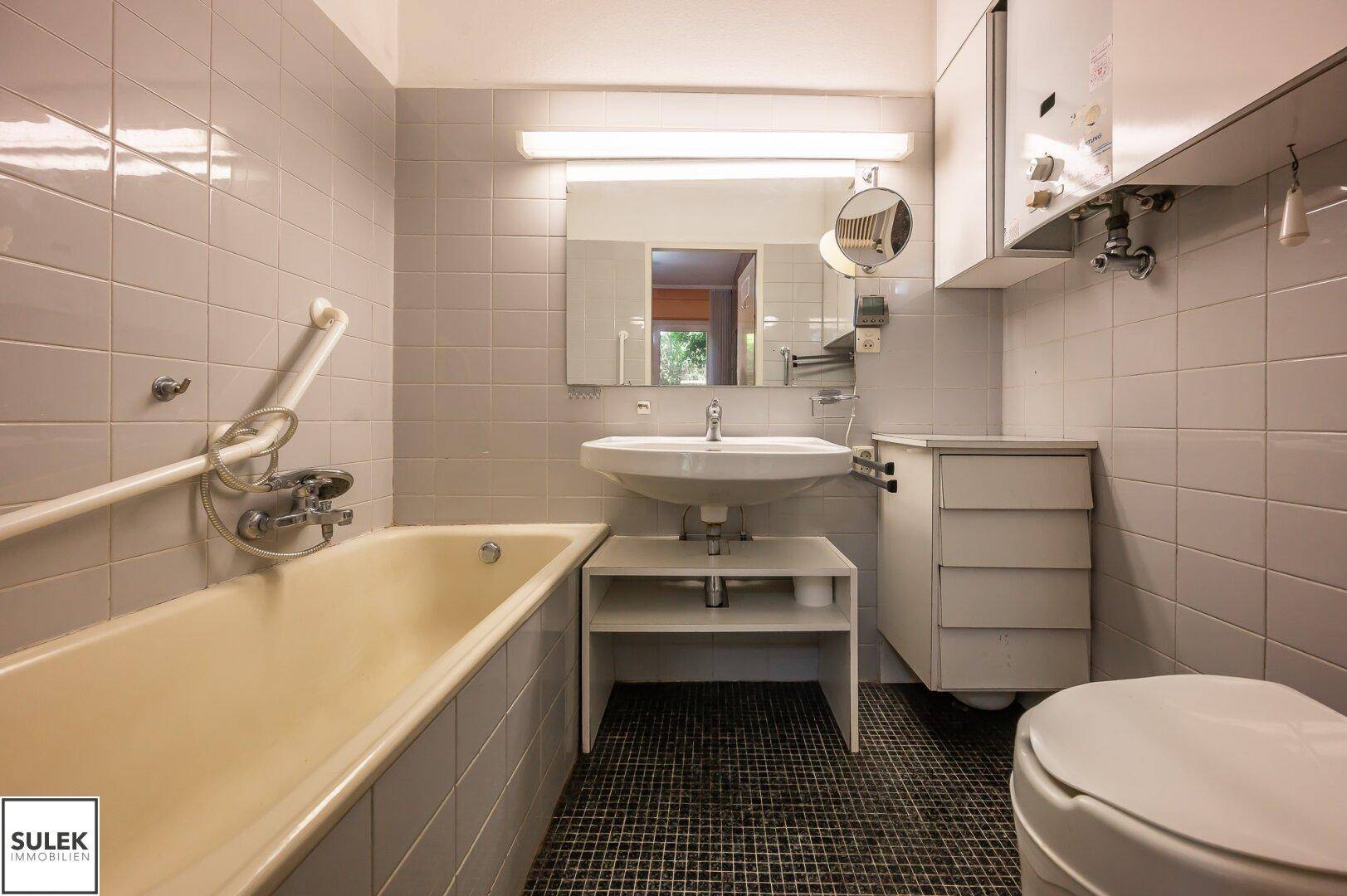 Ein Badezimmer mit Badewanne, Waschbecken und WC, ausgestattet mit hellgrauen Fliesen und Haltegriffen.