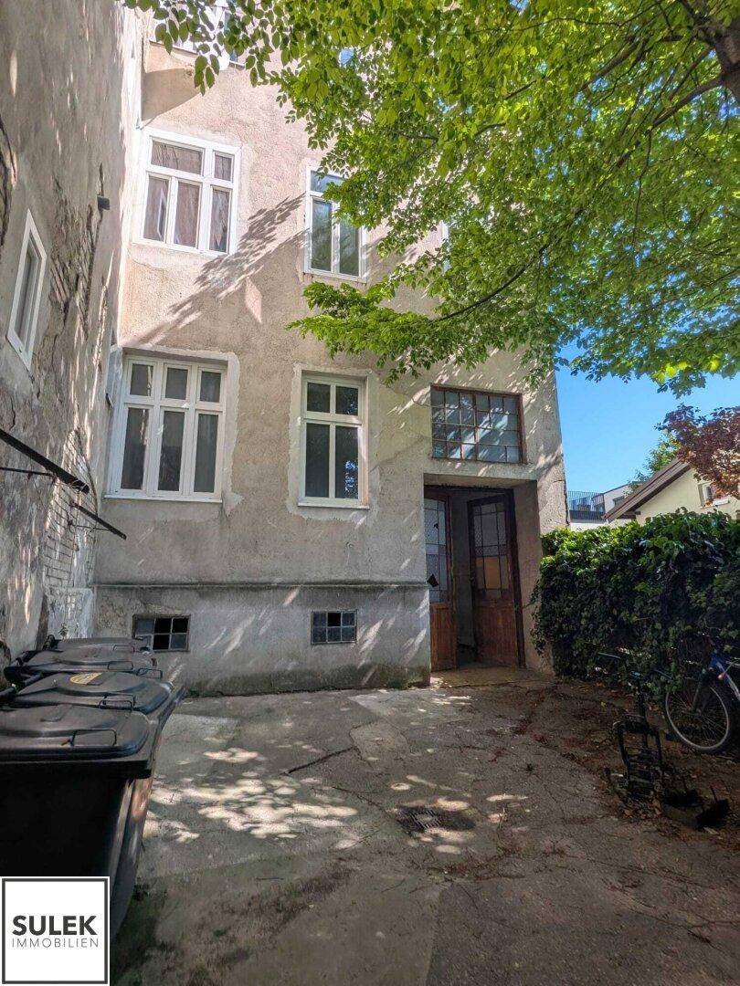 Hinterhofbereich eines Gebäudes mit Mülltonnen und einem großen Baum.