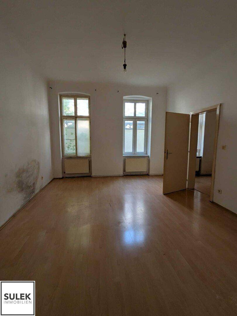 Geräumiger Raum mit Holzfußboden, zwei Fenstern und Renovierungsbedarf an den Wänden.