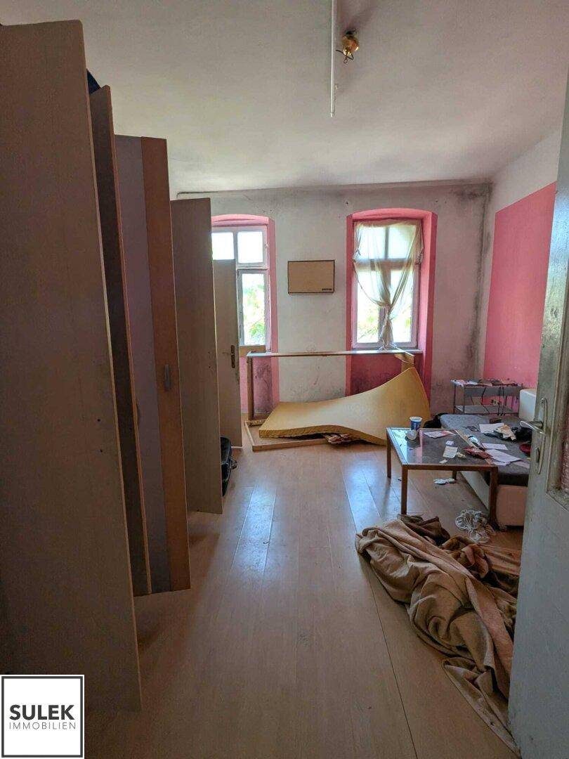 Zimmer mit Holzfußboden, farbigen Wänden und zwei Fenstern, das renoviert werden muss.