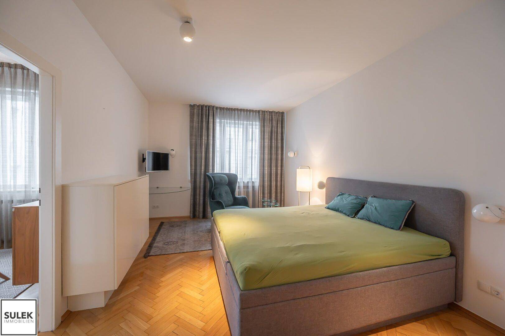 Helles Schlafzimmer mit Doppelbett, Parkettboden und einem Sessel am Fenster.