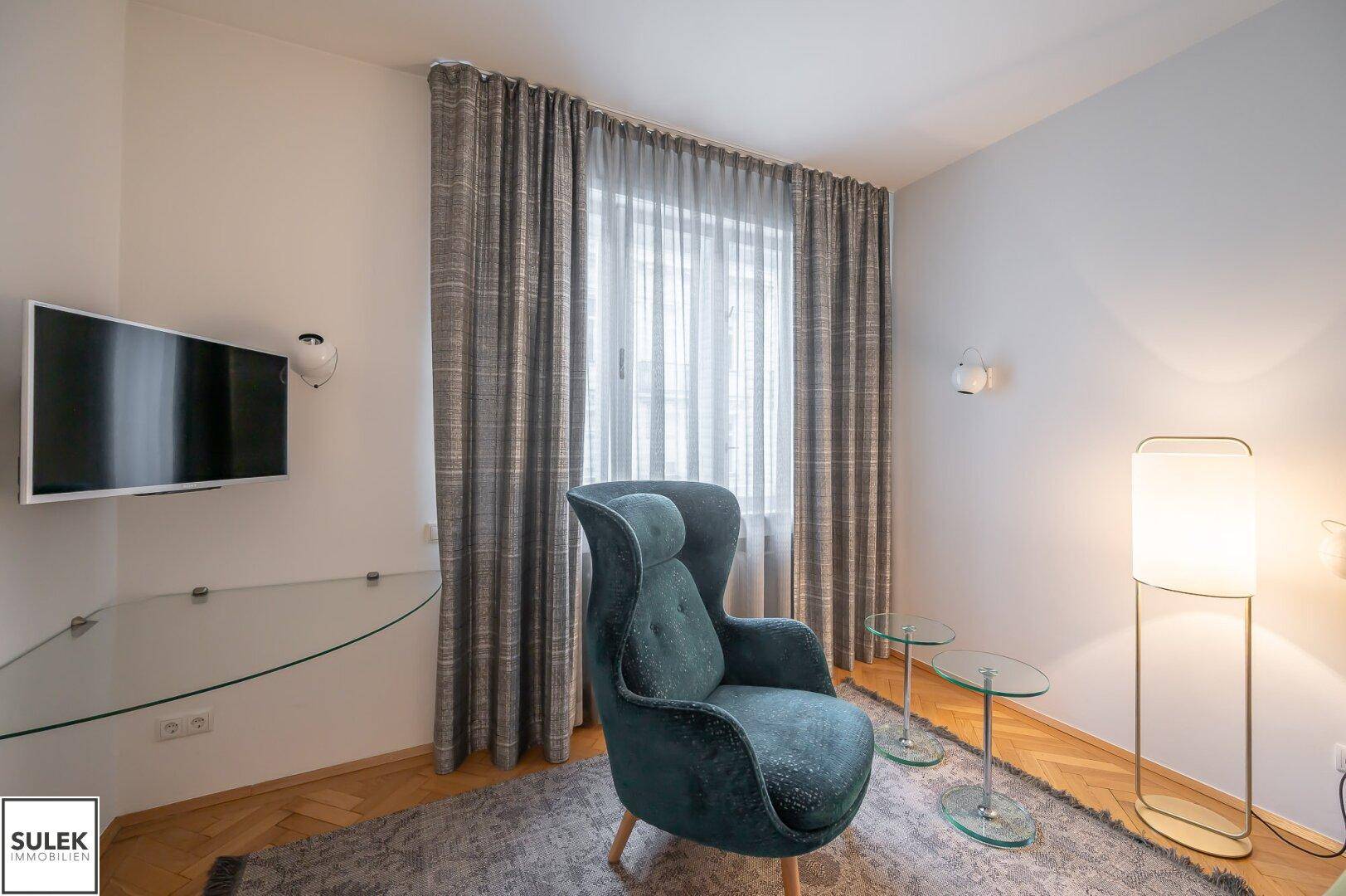 Gemütliche Sitzecke im Schlafzimmer mit Sessel, Fernseher und großen Fenstern.