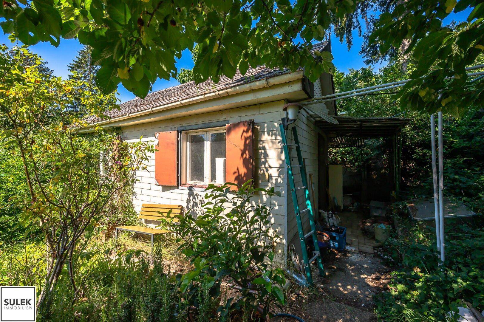 Kleines, älteres Haus oder Gartenhaus mit Holzfensterläden, umgeben von üppigem Grün und einer Bank.