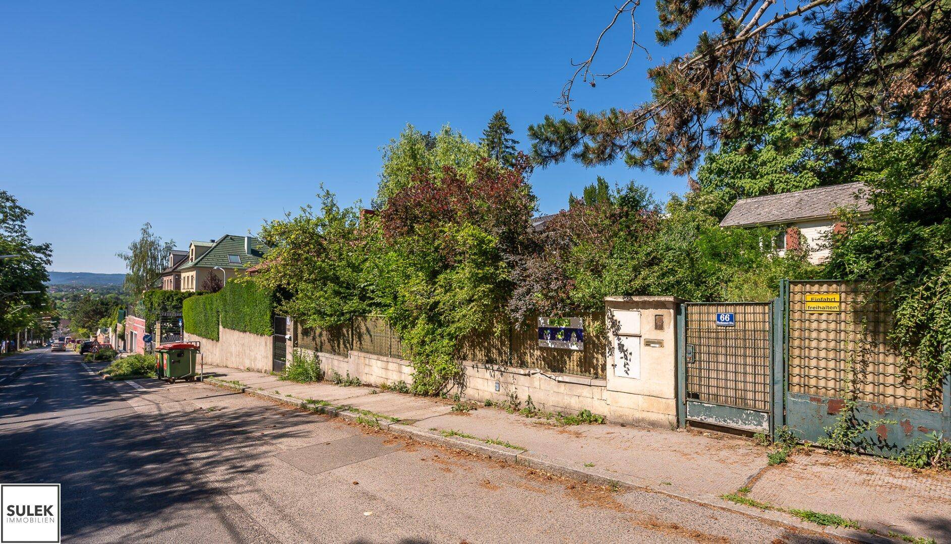 Straßenansicht mit einem Zaun, Tor und Häusern im Hintergrund, umgeben von viel Grün und Bäumen.