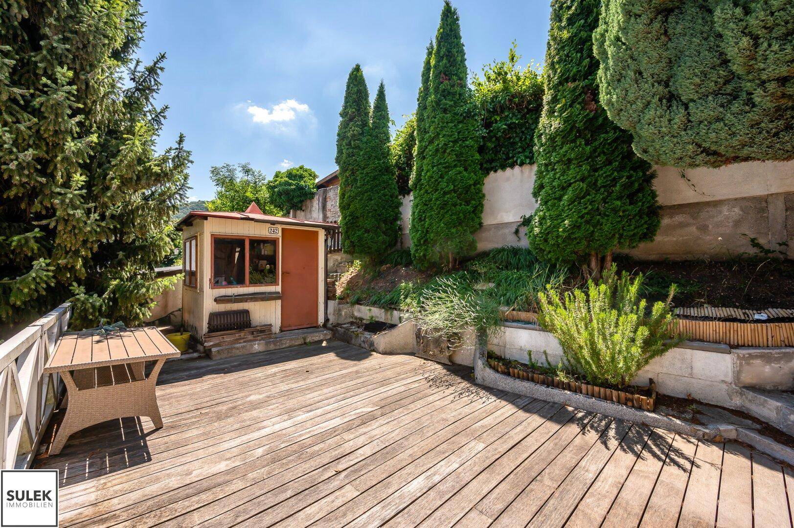 Geräumige Holzterrasse mit Gartenhaus und üppiger Bepflanzung im Hintergrund.