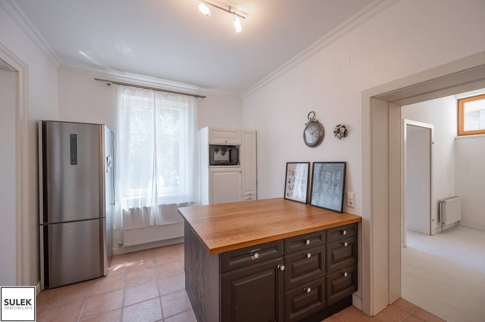 Helle Küche mit Kücheninsel, Kühlschrank und Fenster, das viel Tageslicht hereinlässt.