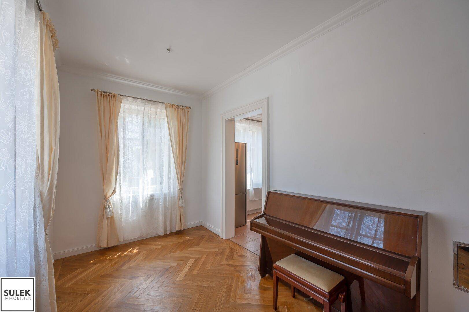Gemütliches Wohnzimmer mit Fischgrätparkett, großem Fenster und Klavier.