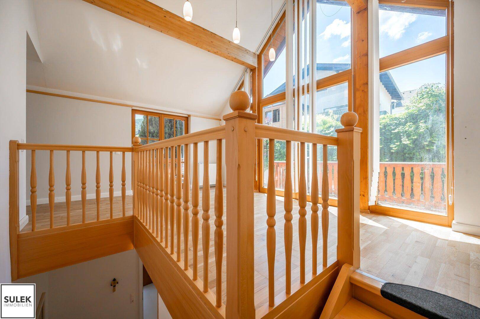 Heller Wohnbereich mit Holztreppe, sichtbaren Holzbalken und großen Fenstern mit Blick ins Grüne.