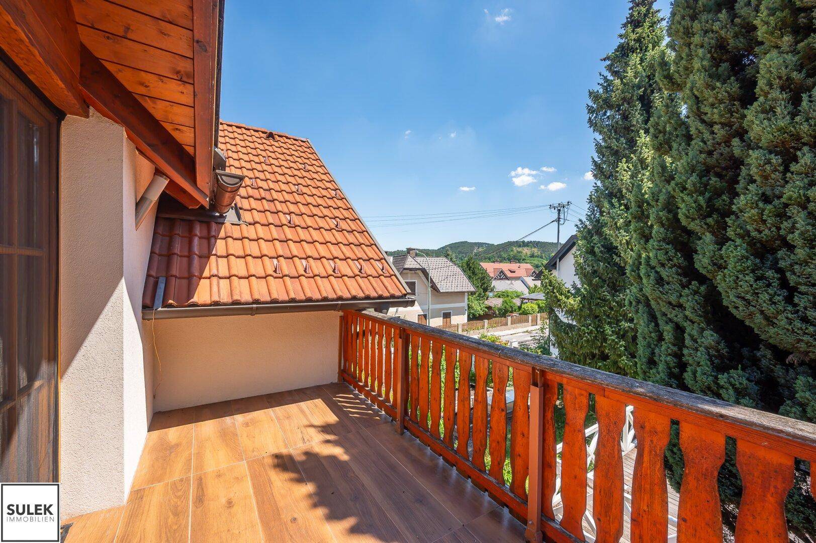 Sonniger Holzbalkon mit traditionellem Geländer und weitem Blick über die Dächer und Landschaft.