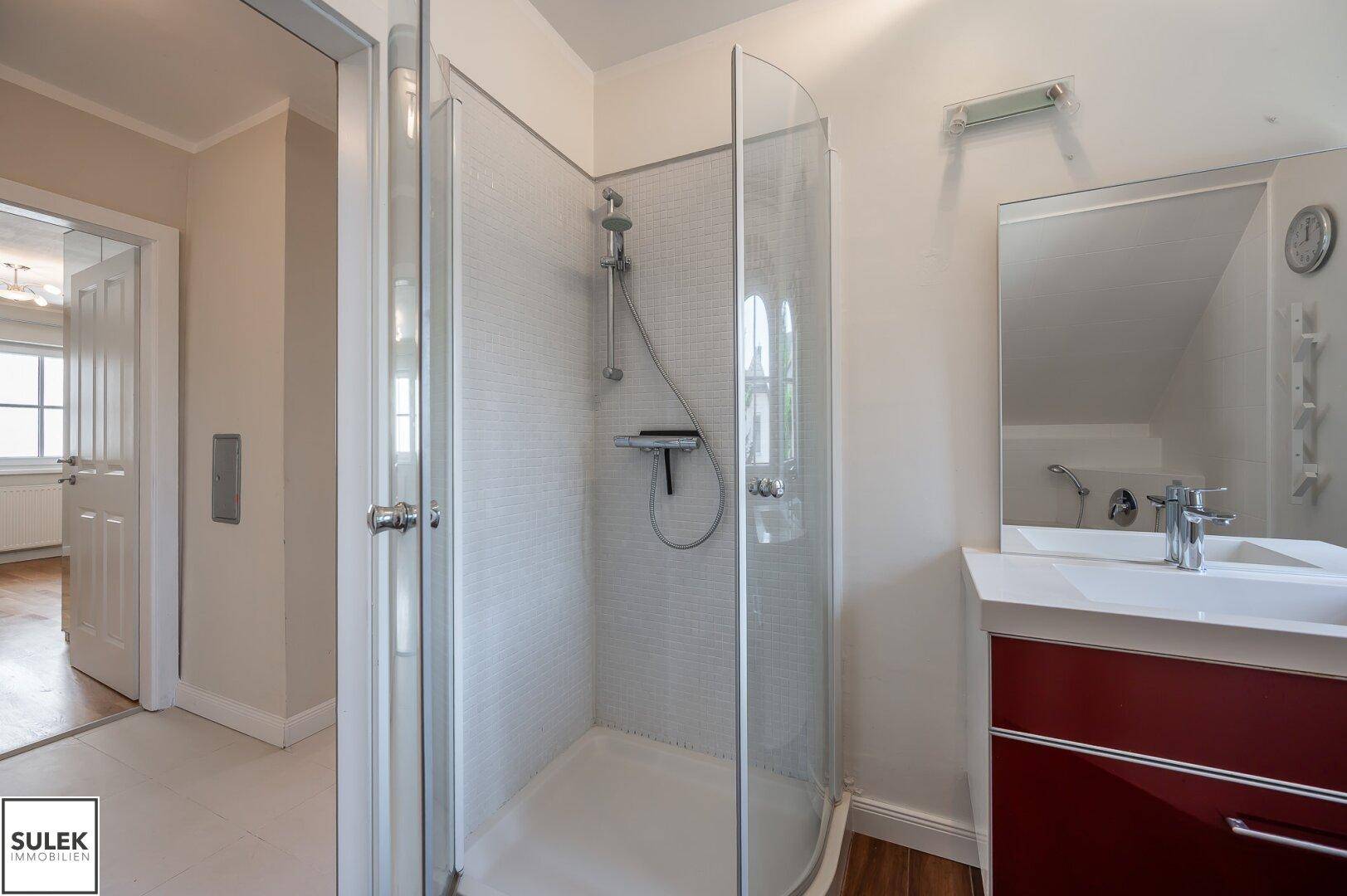 Badezimmer mit Duschkabine, Waschtisch und Spiegel, verbunden mit einem hellen Flur.