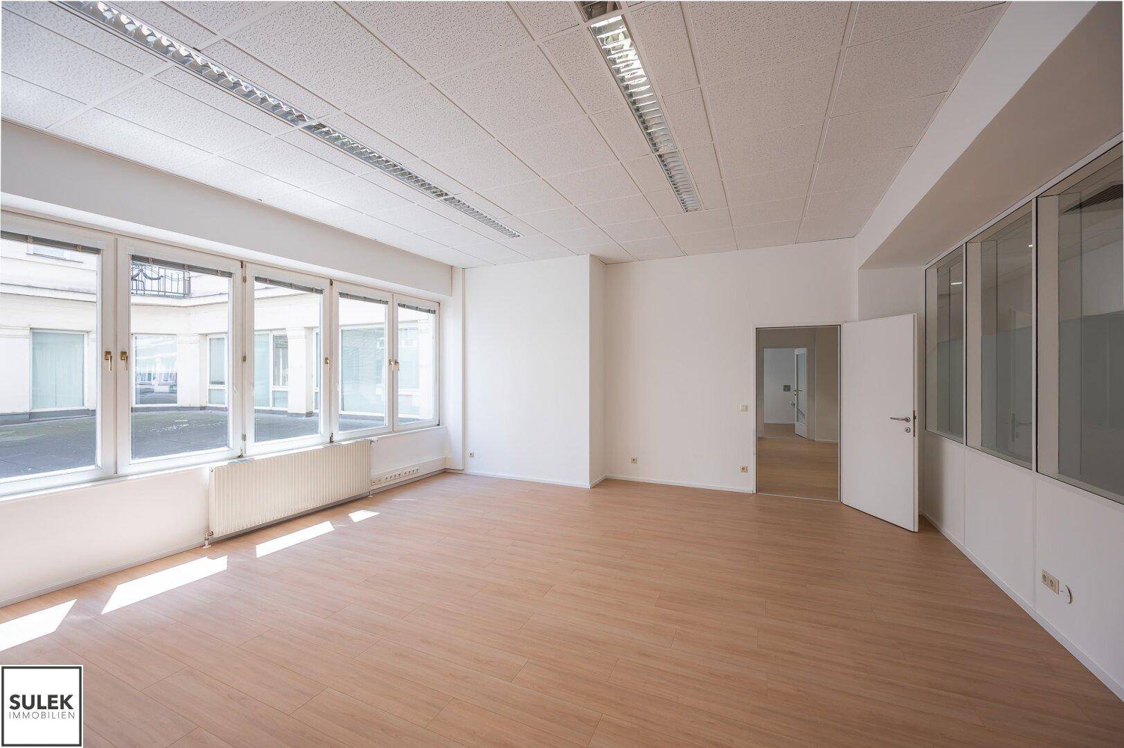 Helles Büro mit großen Fenstern und Blick auf einen Innenhof, ausgestattet mit hellem Laminatboden.