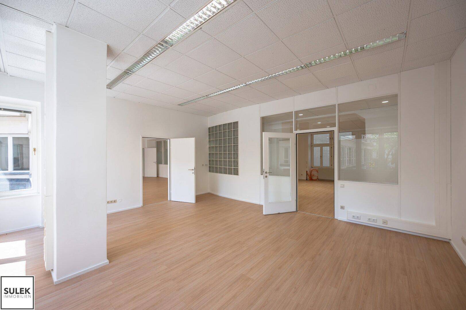 Geräumiges Büro mit hellem Laminatboden, Glaswänden und Türen, die zu weiteren Räumen führen.