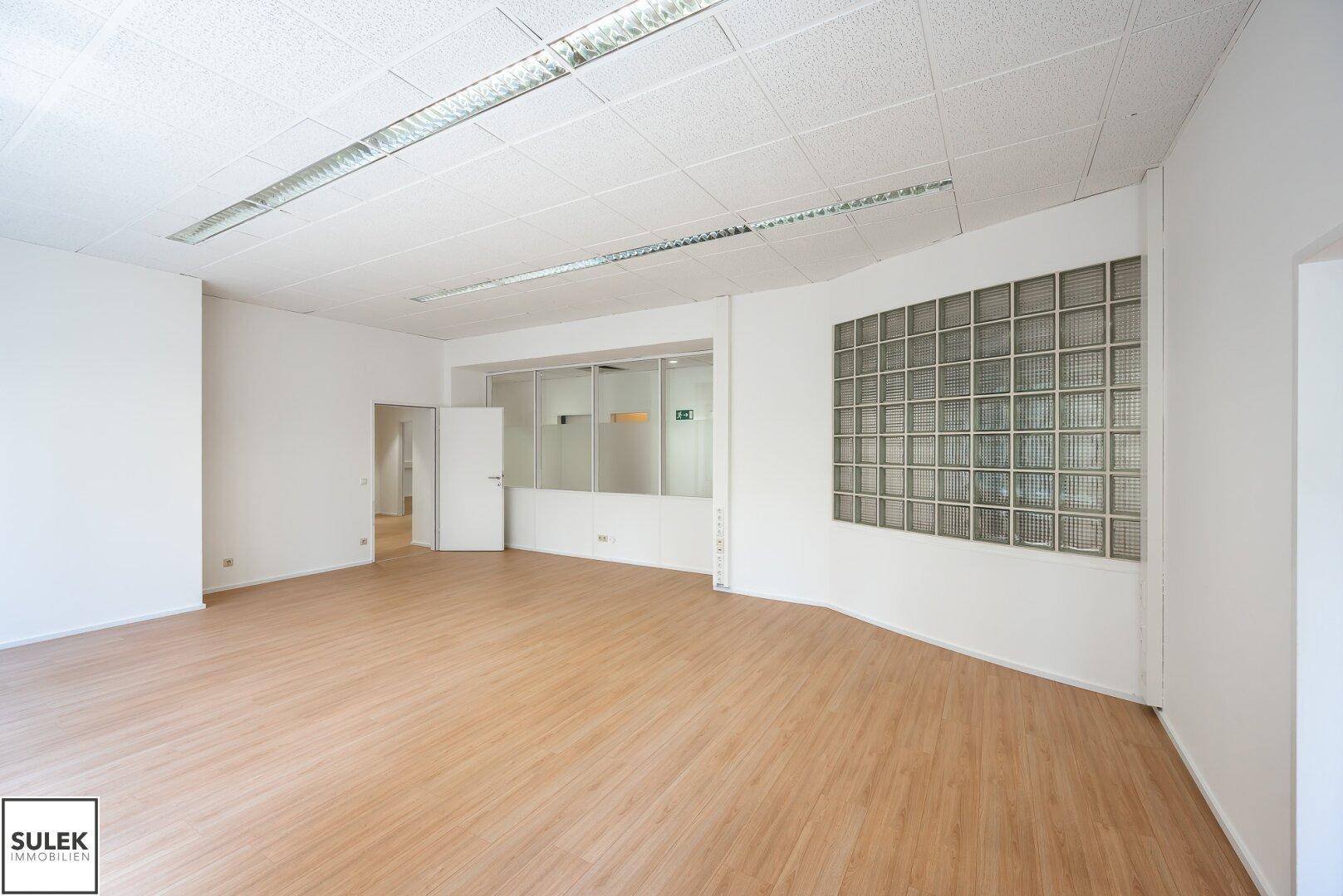 Helles Büro mit Glasbausteinwand und hellem Laminatboden, das zu weiteren Räumen führt.