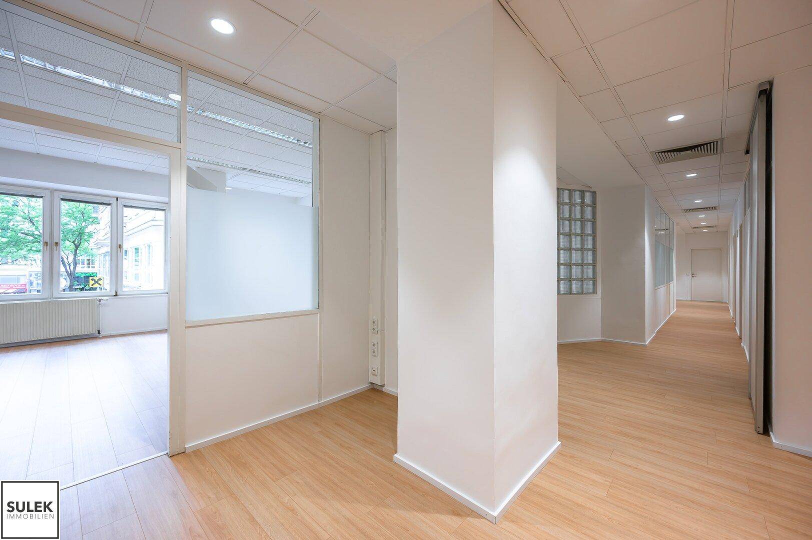 Geräumiger Bürogang mit hellem Laminatboden und Blick in ein angrenzendes Büro mit Fenstern.
