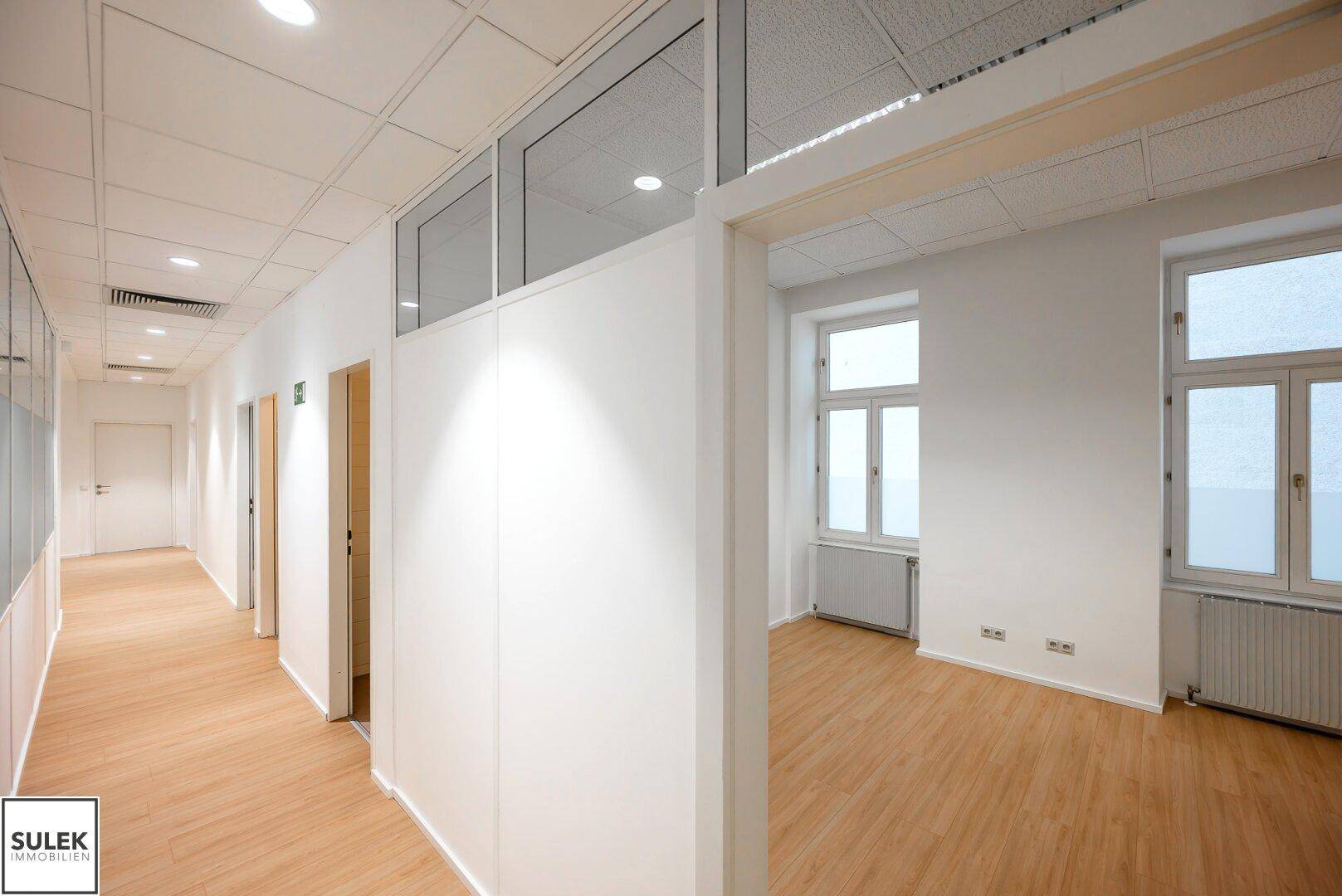Heller Bürogang mit Glaswänden und Türen, die zu verschiedenen Büroräumen führen.