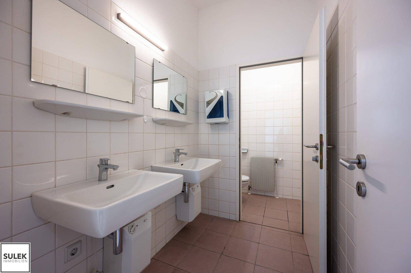 Heller Sanitärraum mit zwei Waschbecken, großen Spiegeln und weißen Fliesen an den Wänden.
