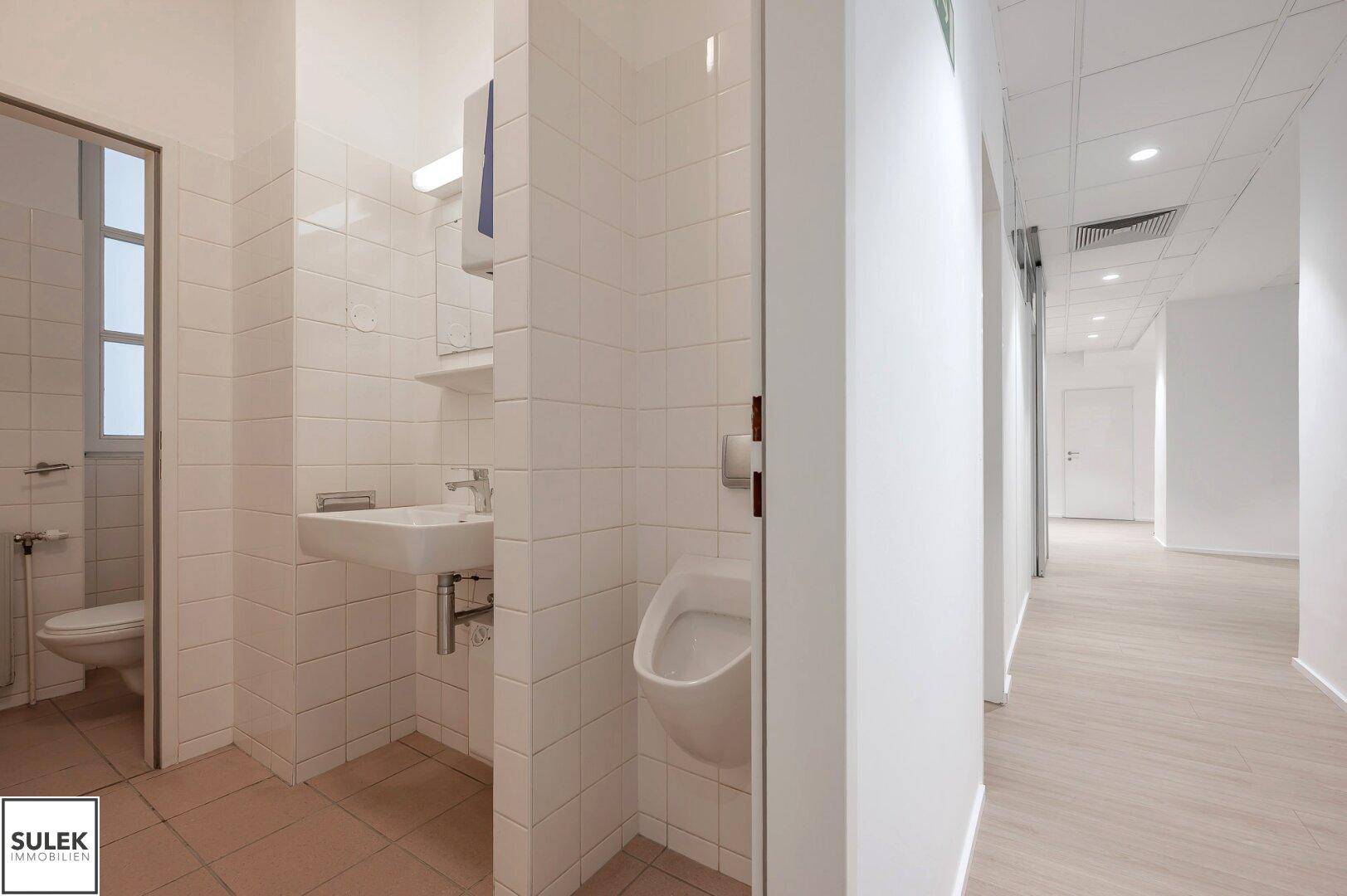 Sanitärbereich mit Urinal, Waschbecken und gefliesten Wänden, angrenzend an einen Bürogang.
