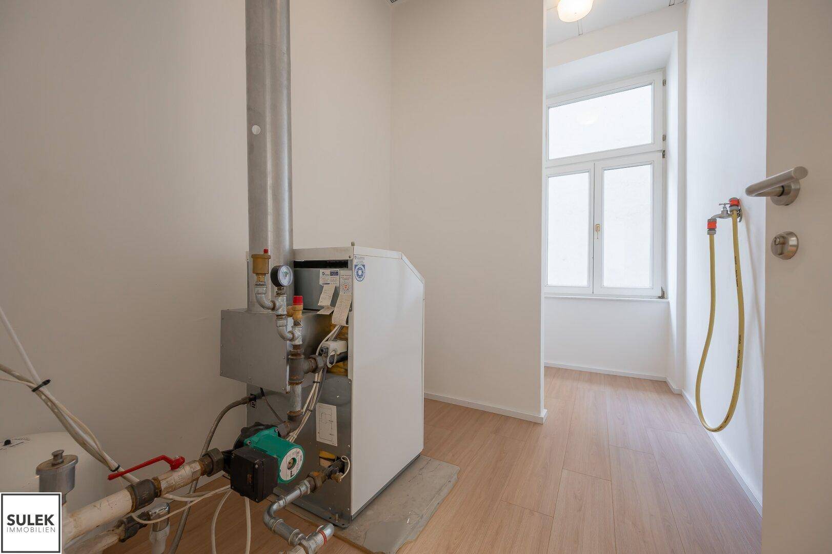 Heizungsraum mit moderner Heizungsanlage, hellen Wänden, Laminatboden und einem Fenster, das Tageslicht hereinlässt.