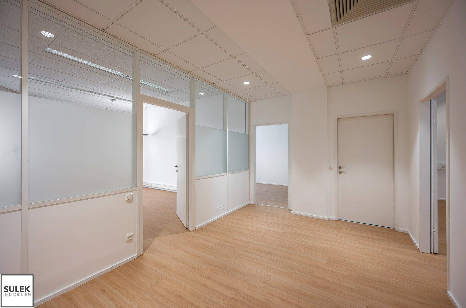 Geräumiges Büro mit hellem Laminatboden, Glaswänden und Türen, die zu weiteren Räumen führen.