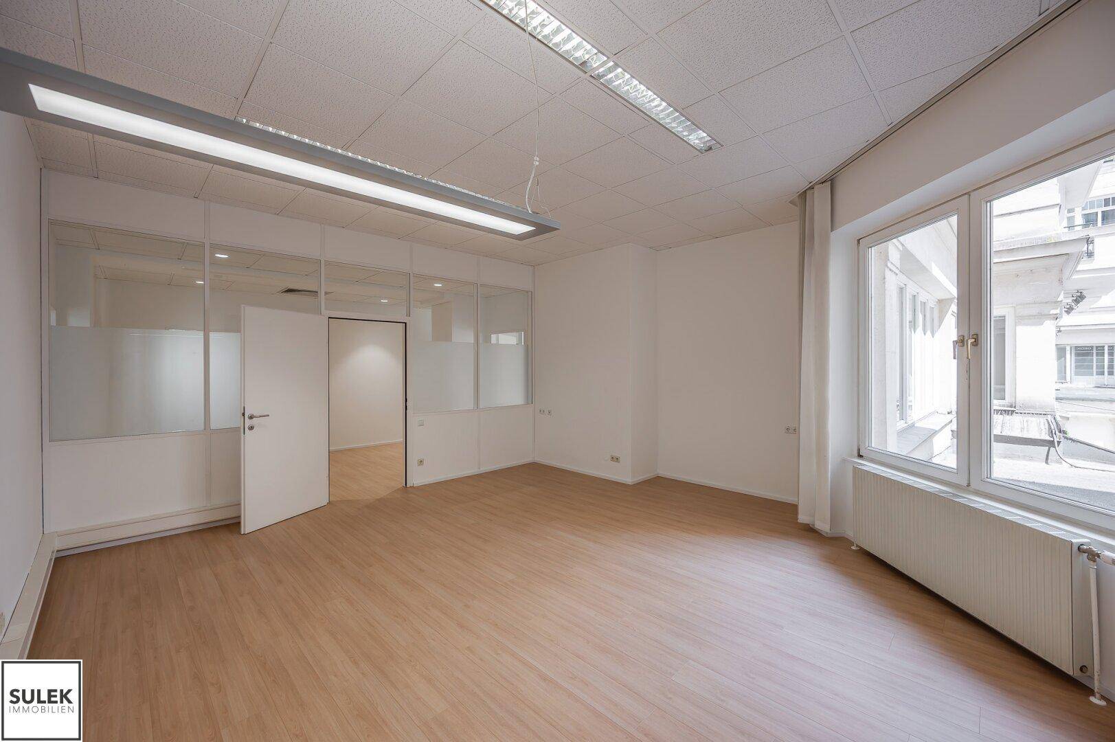 Geräumiges Büro mit großen Fenstern, hellem Laminatboden und abgehängter Decke mit Leuchtstoffröhren.