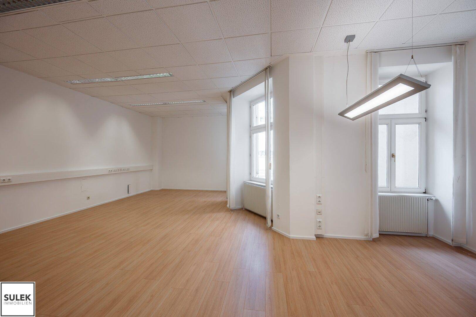 Helles Büro mit zwei großen Fenstern, die viel Tageslicht hereinlassen, und einer modernen Hängelampe.