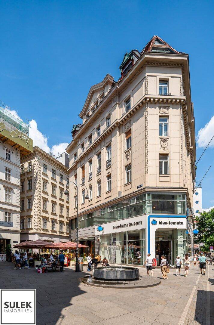 Historisches Gebäude mit Geschäften im Erdgeschoss, einem Brunnen und belebter Straße.