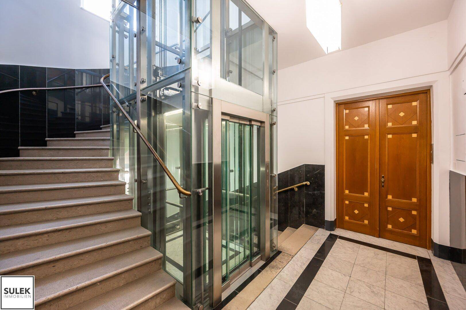 Repräsentativer Eingangsbereich mit einem modernen Glasaufzug und einer eleganten Steintreppe.