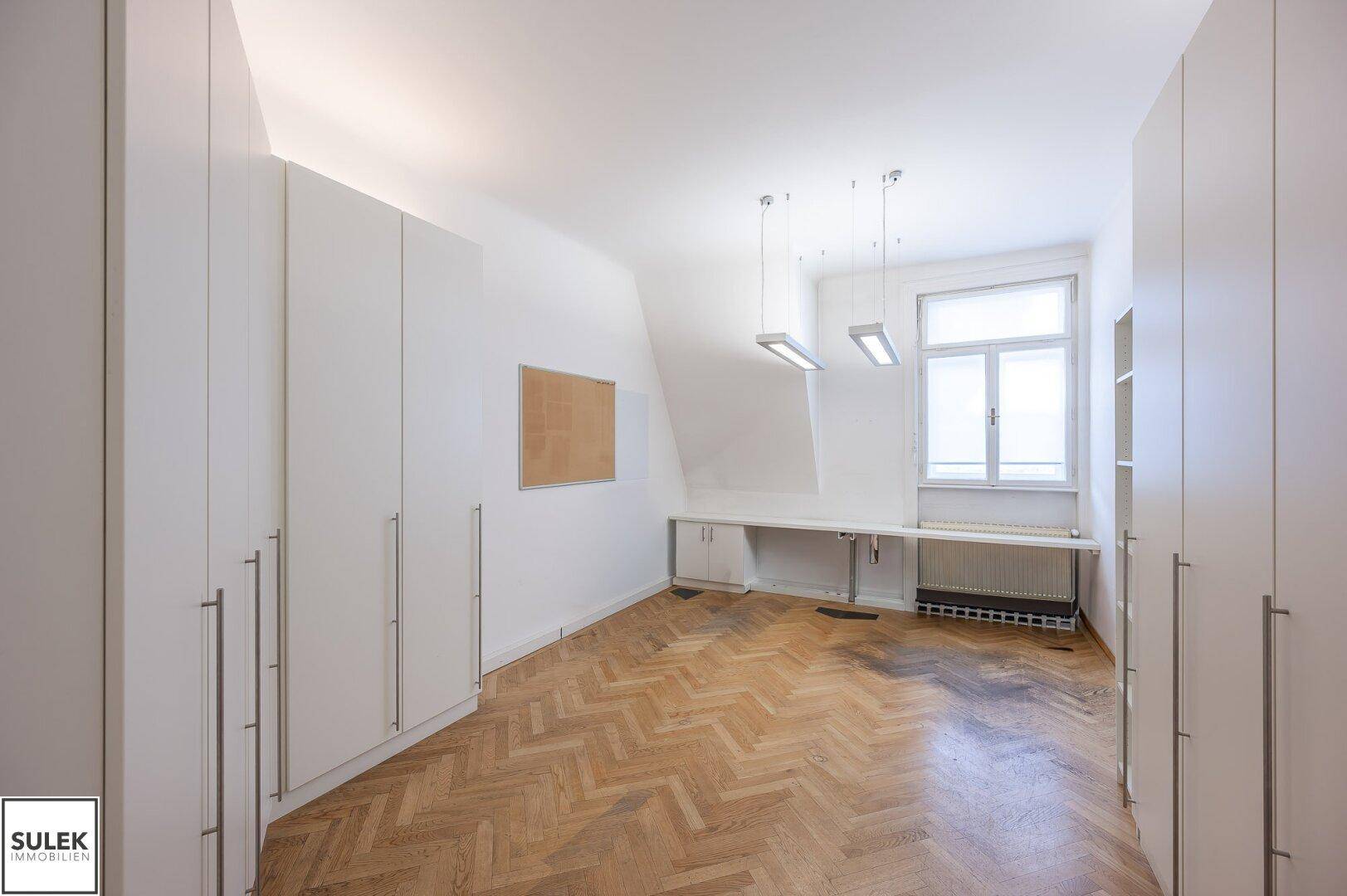 Büro mit Fischgrätparkett, Einbauschränken und einem Fenster mit Blick nach draußen.