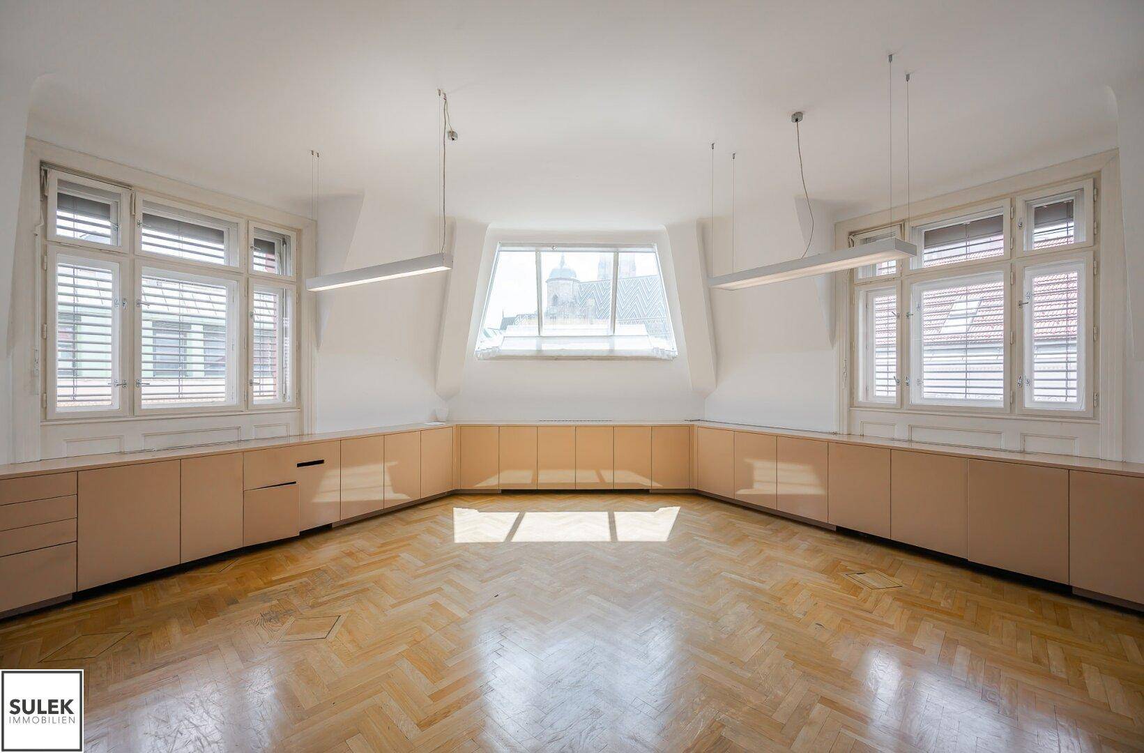 Helles Büro mit Fischgrätparkett, vielen Fenstern und einer langen Reihe von Einbauschränken.