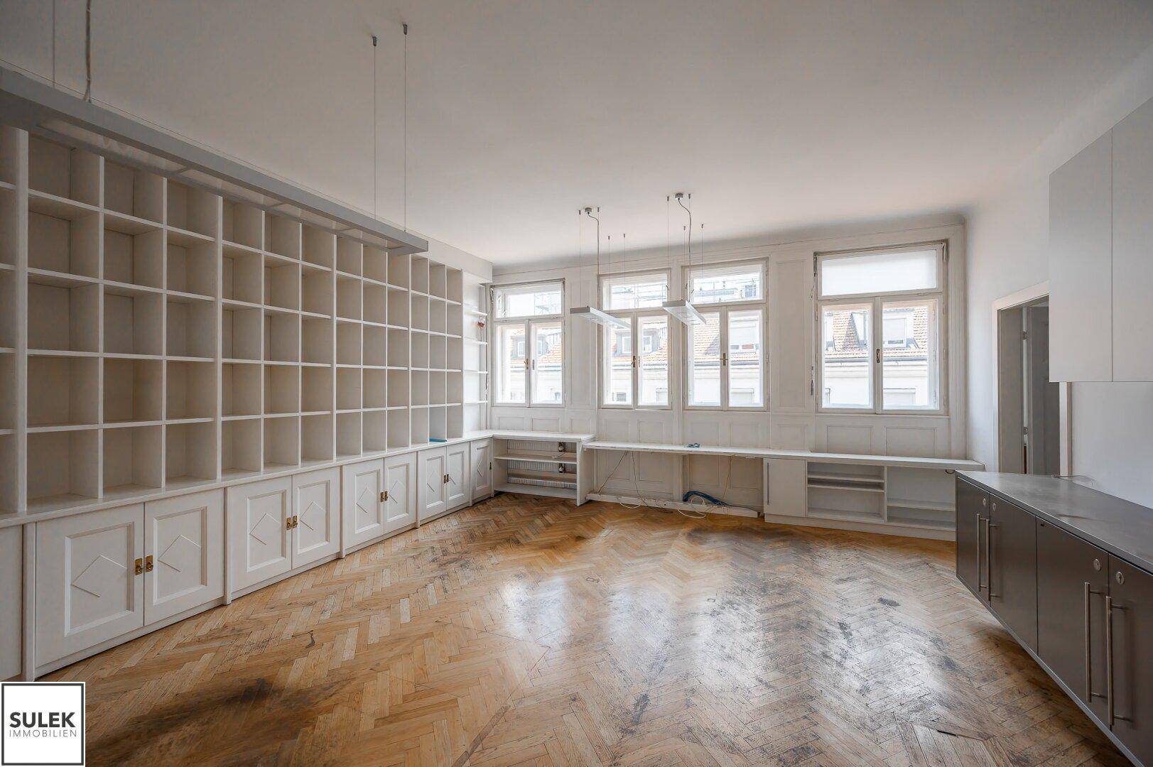 Großes Büro mit Fischgrätparkett, vielen Fenstern und umfangreichen Einbauregalen und Schränken.