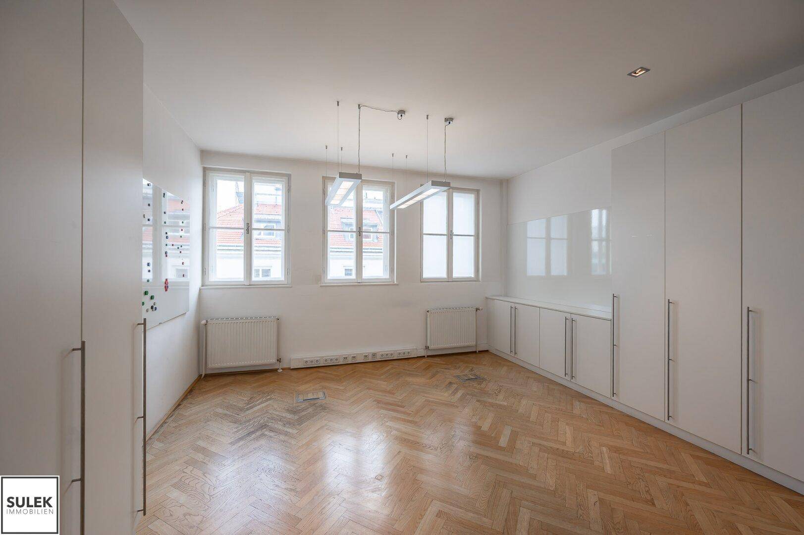 Helles Büro mit Fischgrätparkett, Fenstern und weißen Einbauschränken entlang der Wände.