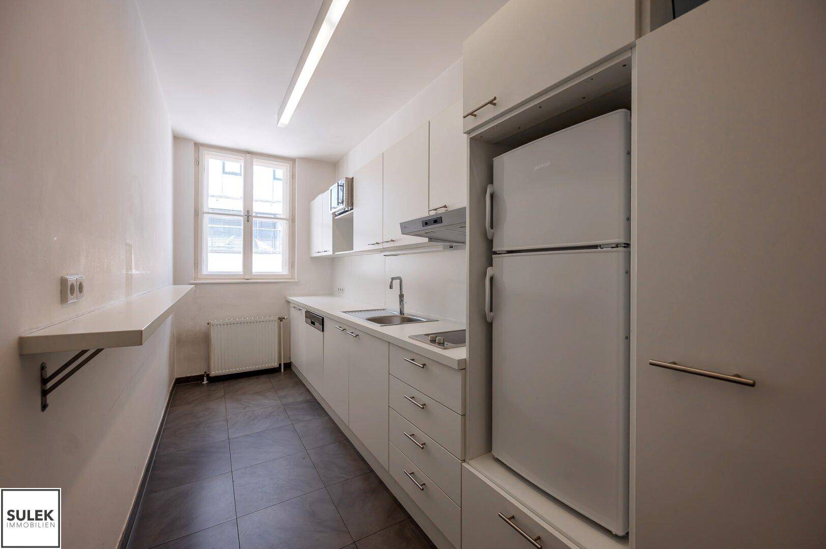Schmale Küche mit weißen Einbauschränken, Spüle, Kühlschrank und Fenster.