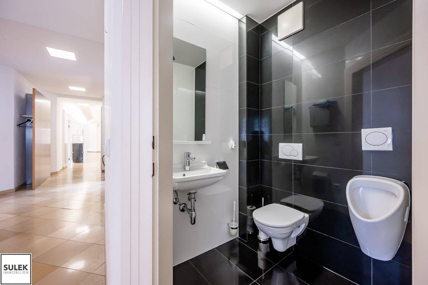 Helles Badezimmer mit schwarzen Fliesen, WC, Urinal und Blick in den angrenzenden Flur.