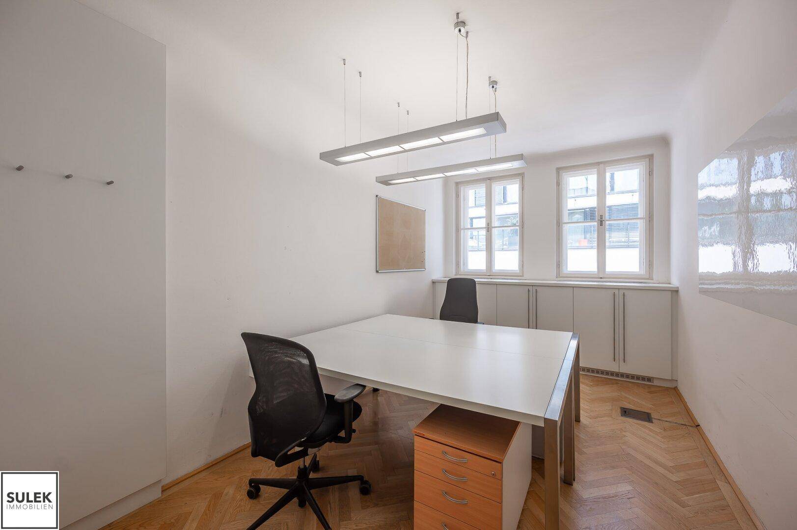 Büro mit Schreibtisch, Bürostühlen, Fischgrätparkett und Fenstern mit natürlichem Licht.