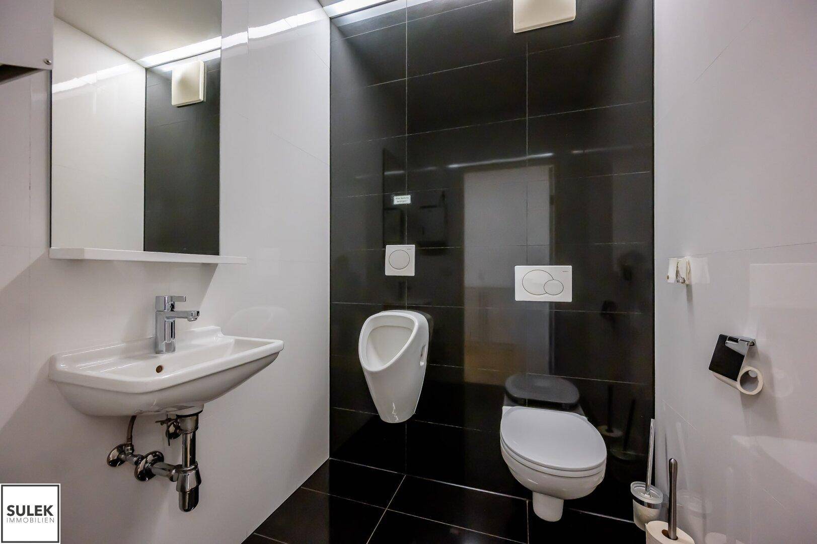 Modernes Badezimmer mit schwarzen und weißen Fliesen, ausgestattet mit WC, Urinal und Waschbecken.