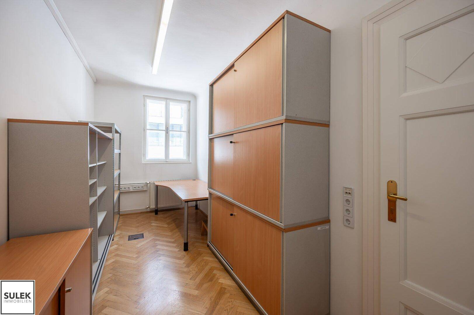 Büro mit Fischgrätparkett, Fenster und modularen Holzschränken für Stauraum.
