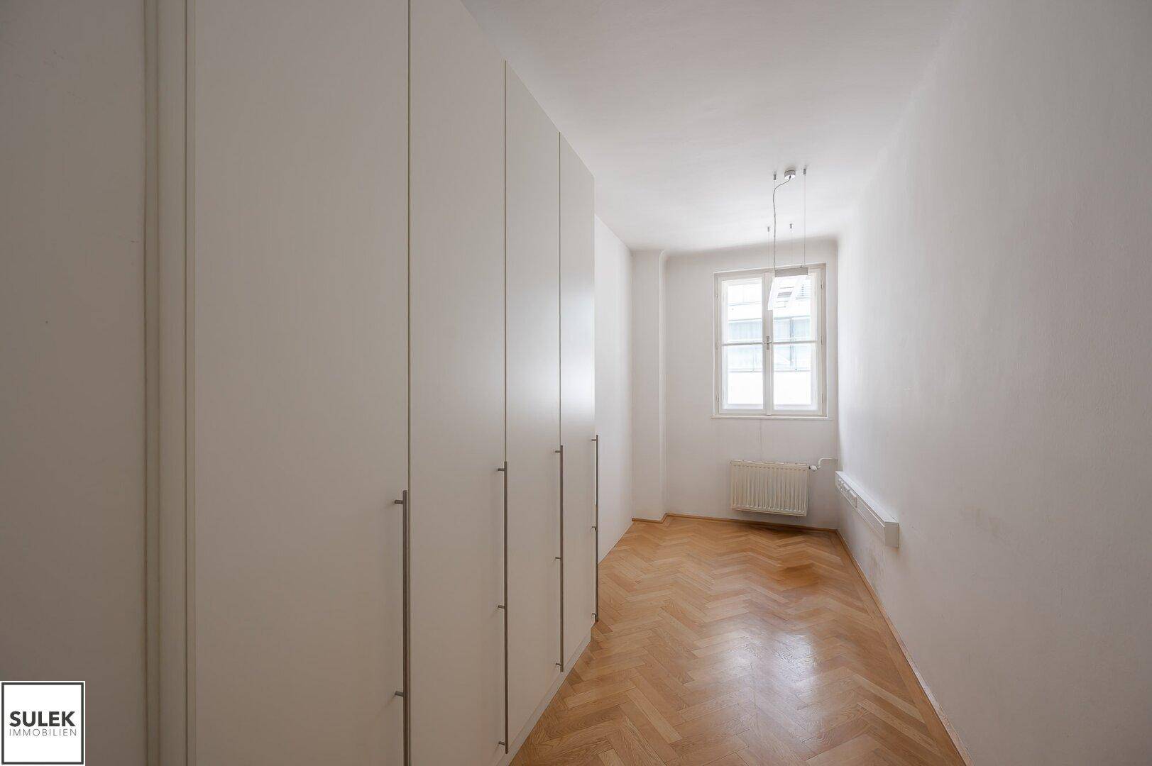 Schmaler Büroraum mit Fischgrätparkett, Fenster und hohen weißen Einbauschränken.
