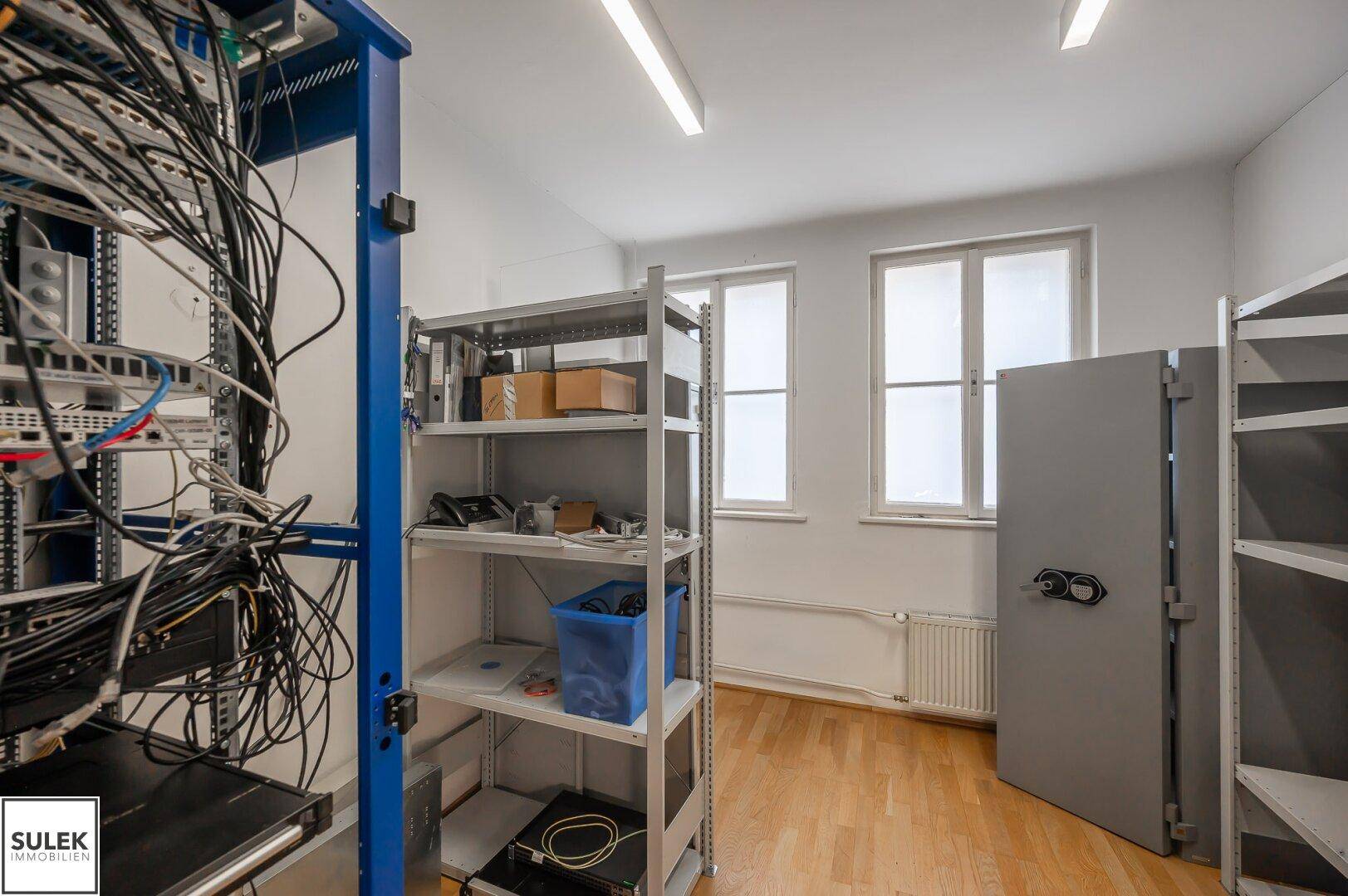 Technikraum mit Serverracks, Regalen und einem großen Safe, beleuchtet durch Fenster.