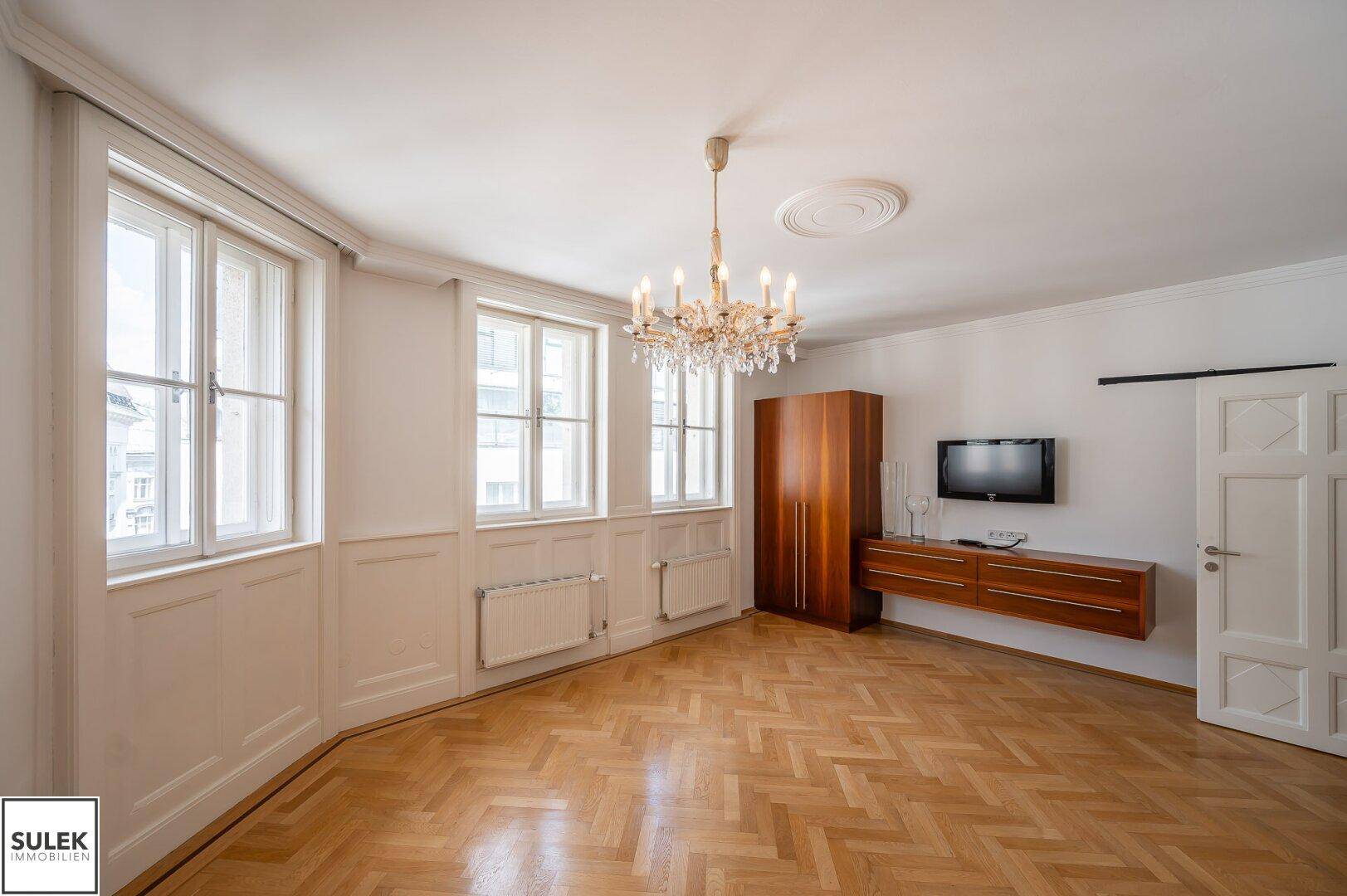 Eleganter Raum mit Fischgrätparkett, Kronleuchter, Wandpaneelen und einem Fernseher an der Wand.