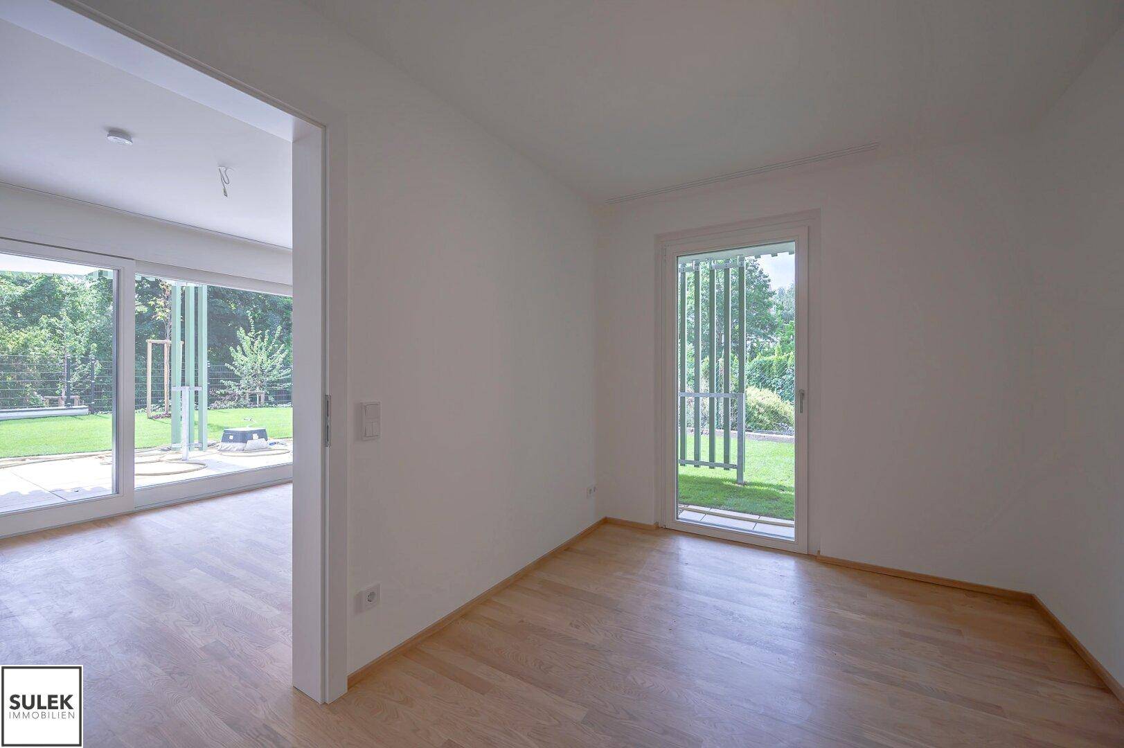 Helles Zimmer mit Holzboden und Zugang zu einem Außenbereich mit Grünblick.