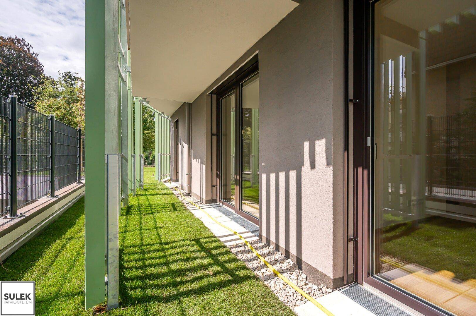 Schmaler Gartenstreifen entlang der Fassade mit großen Fenstern und Zugang zu den Innenräumen.