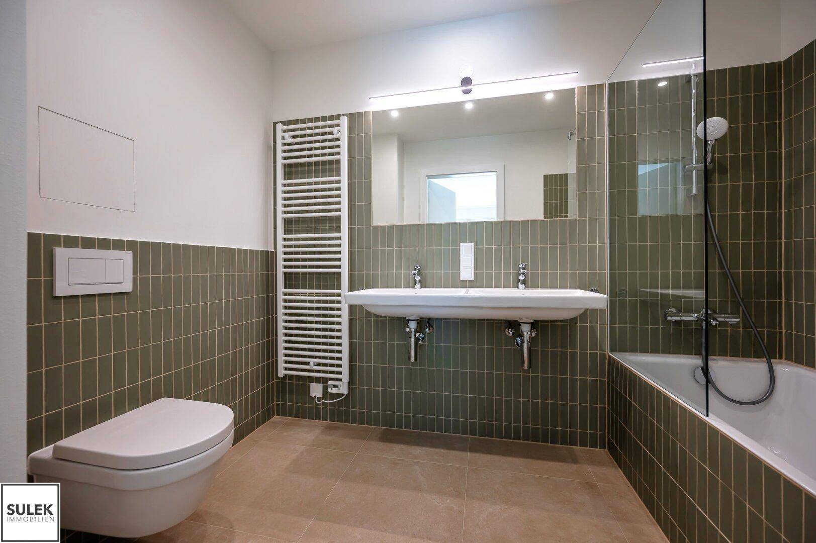 Modernes Badezimmer mit Doppelwaschbecken, Badewanne, WC und grün-grauen Fliesen.