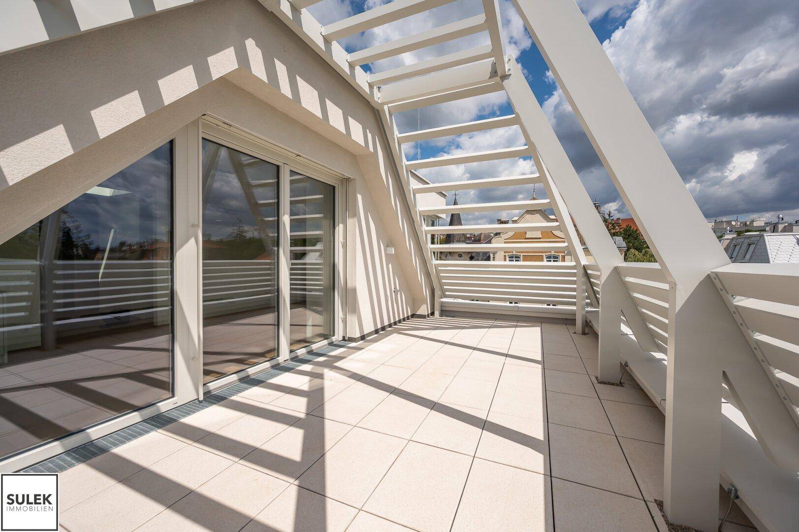 Sonnige Dachterrasse mit Fliesenboden und Blick auf die umliegende Bebauung und den Himmel.