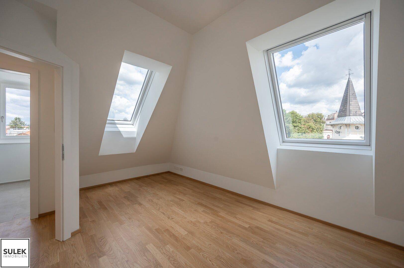 Schlafzimmer im Dachgeschoss mit Holzboden und zwei Dachfenstern, die einen Blick nach draußen bieten.